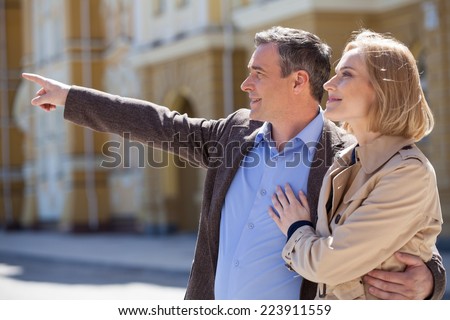 Smiling mature couple standing outside and looking ahead. adult man pointing with finger and hugging woman