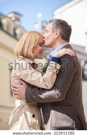 side view of man hugging happy woman outside. Smiling blond woman embracing man outside