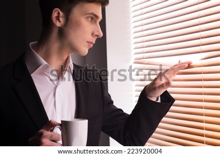 young man looking through window blinds. Handsome business guy peeking through blinds and drinking