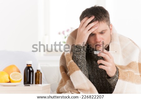 Portrait of sick middle aged man with thermometer. Guy wrapped in plaid sitting on sofa is ill