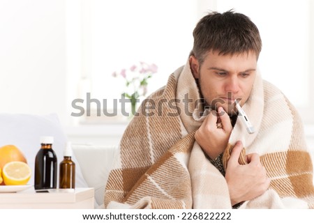 Man with cold sitting on sofa with thermometer in mouth. Man at home sick with flu, taking his temmperature