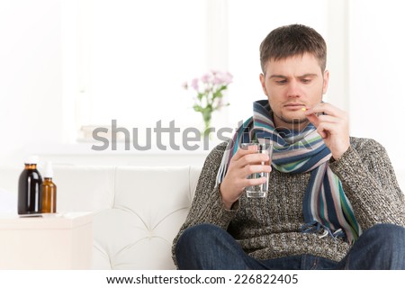 Sick caucasian man holding pills and water. Sick young man with flu drinks water on sofa