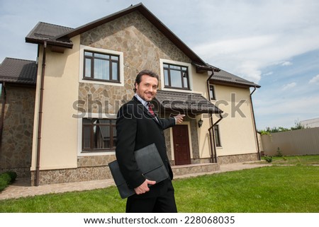 smiling real estate agent ready to sell house. Male real etate agent in front of home pointing to house