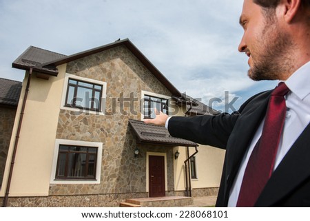 closeup on smiling real estate agent ready to sell house. Male real etate agent in front of home pointing to house window