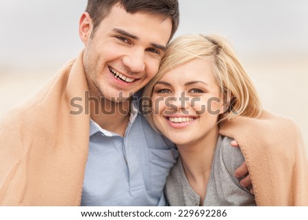 Attractive young couple sitting on beach at summer. happy woman and man sitting under blanket and hugging