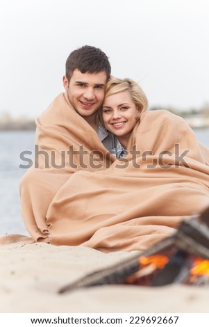 happy couple having fun around bonfire. lovers sitting under blanket and warming near fire