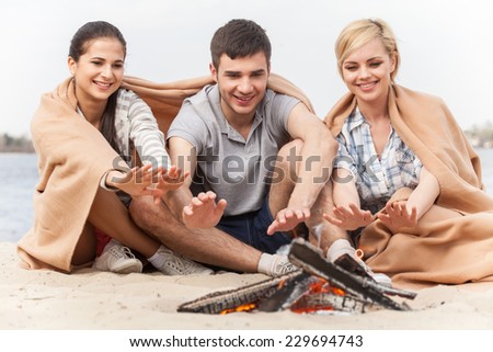 happy friends having fun around bonfire. three friends sitting under blanket and warming near fire