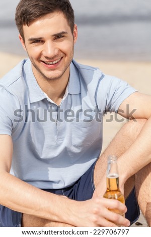 young man holding bottle of beer. smiling guy sitting on beach and looking into camera