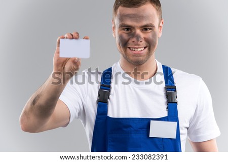 Man in blue overalls holding business card. dirty face of young guy standing on grey background