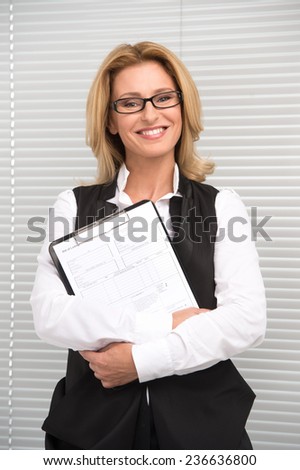 Laughing business woman in white shirt. Attractive mature businesswoman with clipboard before shutters smiling
