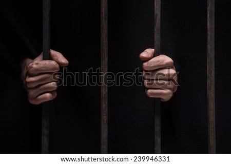 closeup on hands of man sitting in jail. Man behind jail bars on black background