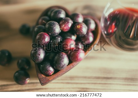 Red wine in crystal stemless glass with wooden heart shaped bowl filled with Muscadine grapes on burned wooden background.
