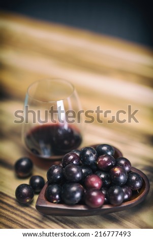 Red wine in crystal stemless glass with wooden heart shaped bowl filled with Muscadine grapes on burned wooden background.