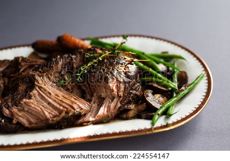 Slow cooked pot roast with carrots, green beans, onions, garlic and gravy on a white porcelain platter with gold rim against a gray tablecloth.