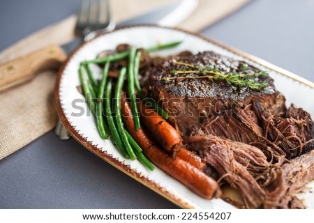 Slow cooked pot roast with carrots, green beans, onions, garlic and gravy on a white porcelain platter with gold rim and serving fork against a gray tablecloth.