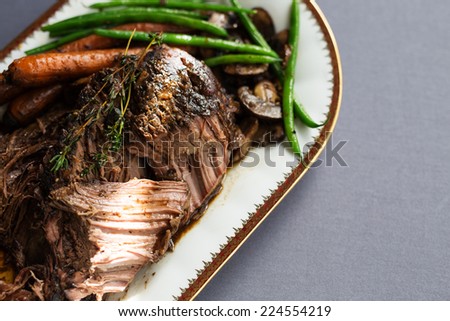 Slow cooked pot roast with carrots, green beans, onions, garlic and gravy on a white porcelain platter with gold rim against a gray tablecloth.