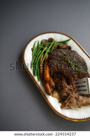 Slow cooked pot roast with carrots, green beans, onions, garlic and gravy on a white porcelain platter with gold rim and serving fork against a gray tablecloth.