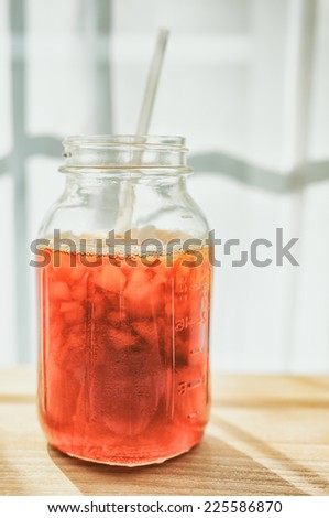 Iced tea in a jar with ice and a straw on a wooden board against a brightly lit background.
