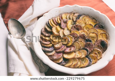 Sliced and roasted potatoes with rosemary and a silver serving spoon in a shallow baking dish. Analog and vintage filters applied.