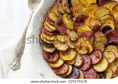 Sliced and roasted potatoes with rosemary and a silver serving spoon in a shallow baking dish.