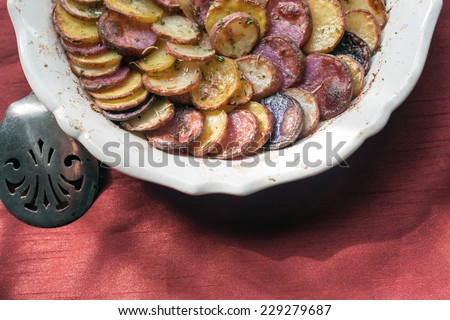 Sliced and roasted potatoes with rosemary and a silver serving spoon in a shallow baking dish on a red silk tablecloth.