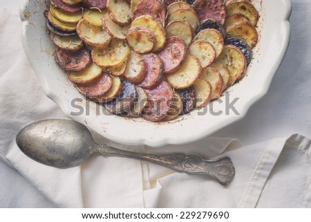 Sliced and roasted potatoes with rosemary and a silver serving spoon in a shallow baking dish. Analog and vintage filters applied.