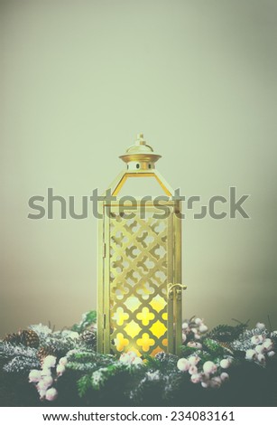 Gold lantern against light green background with a wreath made of pine branches and pine cones.  Faded analog filter.