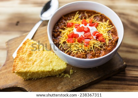 A bowl of chili with tomatoes, sour cream, cheese and a piece of cornbread against a wooden background.