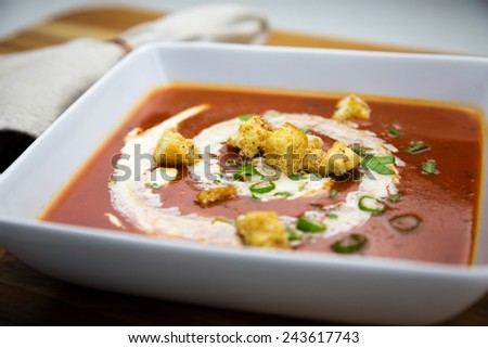 White square bowl filled with tomato soup with cream, scallions and croutons.