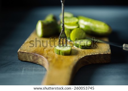 Small silver antique fork through a pickle chip on a cutting board with knife.  Slate gray background.