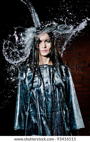 Woman standing under a deluge of water pouring from above her head, upper body pose outside with dark brick wall.