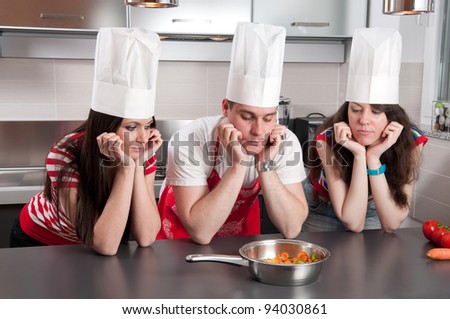 Three chefs with their elbows on the kitchen countertop staring at a pan of carrots with long faces.