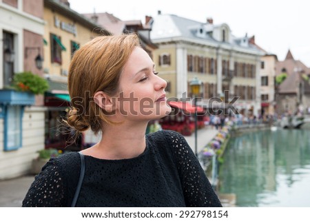 A young woman is standing and smiling. In the background, the French street and the channel.
