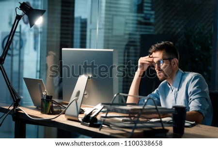 Casual tired office worker sitting at desk using computer and doing overtime project in lamplight.