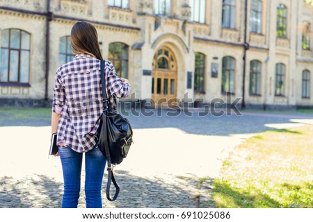 First day at university for young happy female student in casual clothes with backpack on her shoulders. View from back, she is looking at the entrance of the building