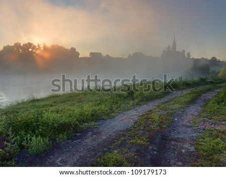 Rural early morning sunrise landscape with lake, road, fog and beautiful sun with rays.