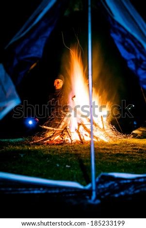 Man near huge camp fire and tent holding lantern and looking at the fire