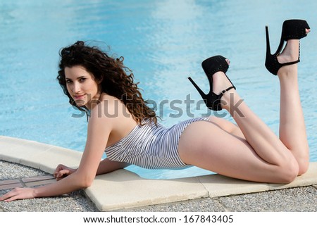 Lady is posing by the pool whilst wearing a one-piece swim suit and high heels