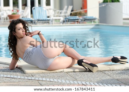 Lady is posing by the pool whilst wearing a one-piece swim suit and high heels - Hotel entrance can be seen in the background