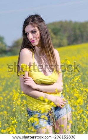 Yellow blooming field, cute fit young lady in yellow shirt. She is hugging herself and holds a bouquet of yellow flowers. She laze about the nice landscape