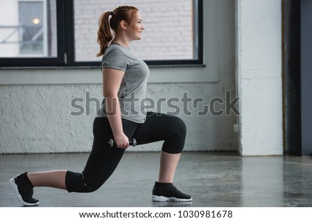 Obese girl performing lunges with dumbbell in gym