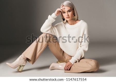 attractive fashionable woman posing in white trendy sweater, beige pants and autumn heels, on grey