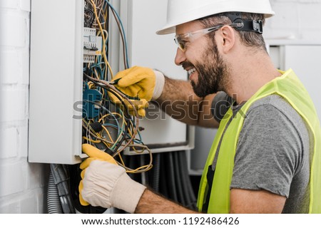 side view of smiling handsome electrician repairing electrical box with pliers in corridor