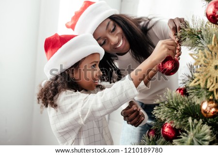 african american mother and daughter in santa claus hats decorating christmass tree together at home