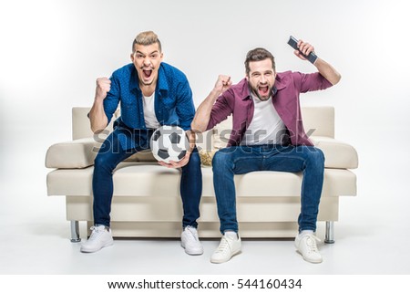 Emotional male friends sitting on couch with soccer ball and supporting favorite team