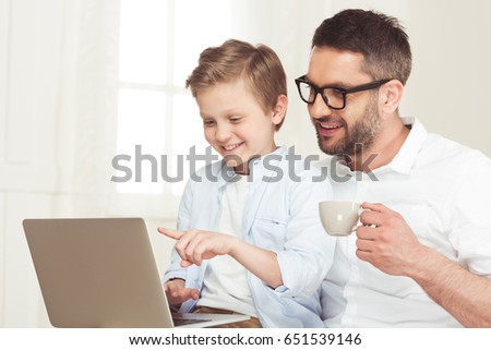 young father drinking coffee and using laptop while with little son at home