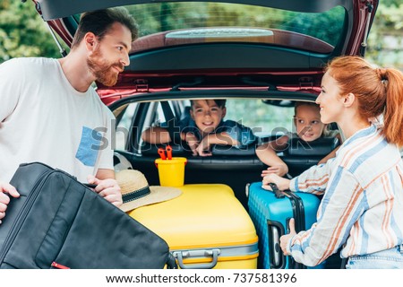 young parents packing luggage in trunk of car with kids on backseats