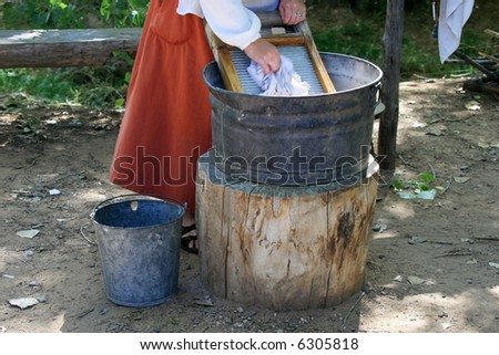 A woman in a long red skirt is washing clothes on a washboard and bucket set up by a stream.  Motion blur on hand scrubbing cloth on the board.