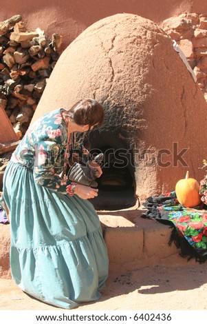 A woman dressed in vintage clothing is baking bread in an outside adobe oven called a horno.