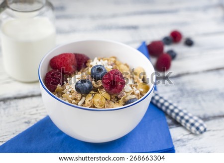close up bowl of muesli breakfast and raspberries and blueberries on bright blue napkin and bottle of milk